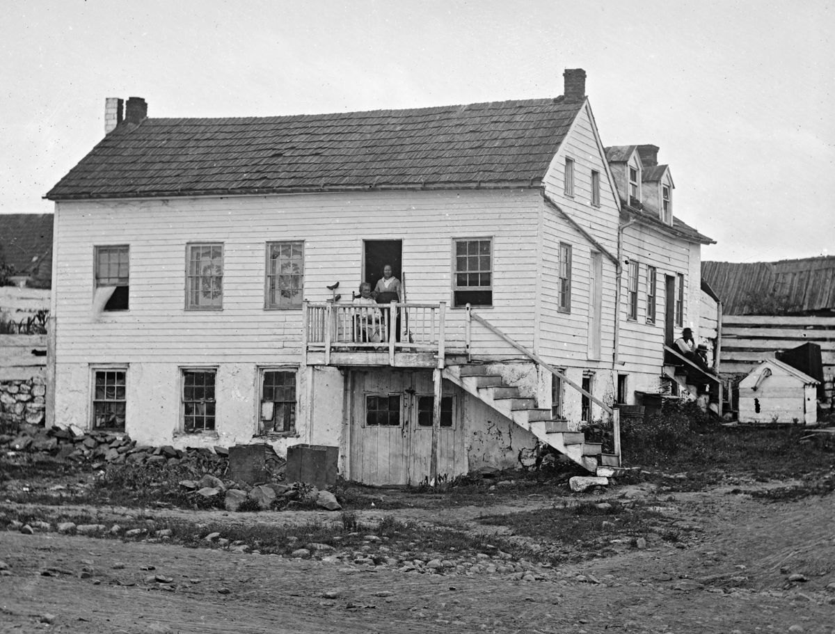 This Brady image, taken from half of a stereo negative, shows citizen hero John Burns sitting on the porch of his house. At right, sitting on the steps is the straw-hatted Brady. To his right, mounted on a tripod behind the water well, is Brady’s portable darkroom.
