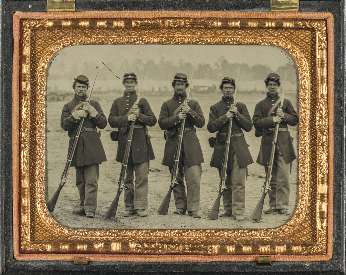 Five soldiers, four unidentified, in Union uniforms of the 6th Regiment Massachusetts Volunteer Militia outfitted with Enfield muskets in front of encampment.